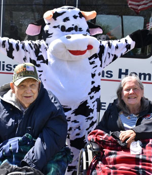 Person in a cow costume standing behind two veterans sitting in lawn chairs.